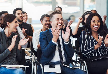 Audience at a conference clapping, with individuals seated in rows and smiling.