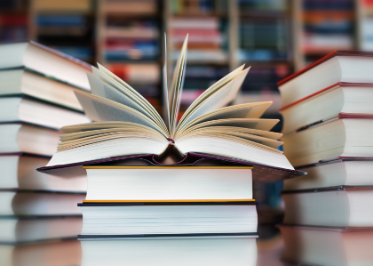 An open book with its pages fanned out, surrounded by stacks of books, with a blurred background of bookshelves filled with more books.