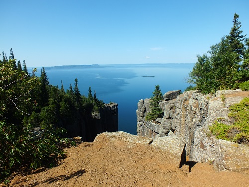 Scenic view of a rocky cliff overlooking a calm, expansive blue lake, surrounded by lush green trees under a clear blue sky.