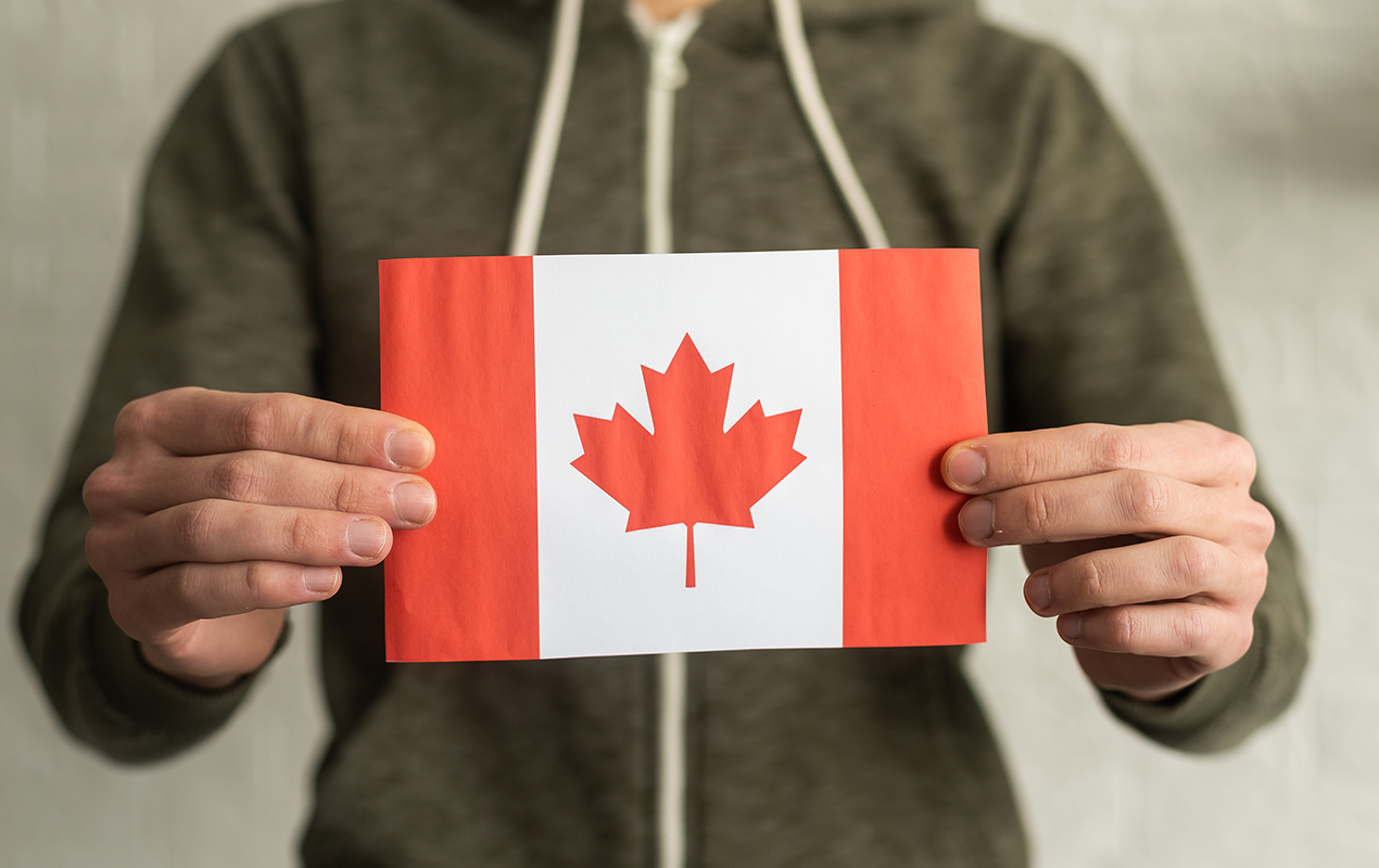 Person holding a small Canadian flag with a red maple leaf on a white background, symbolizing Canada and national pride.