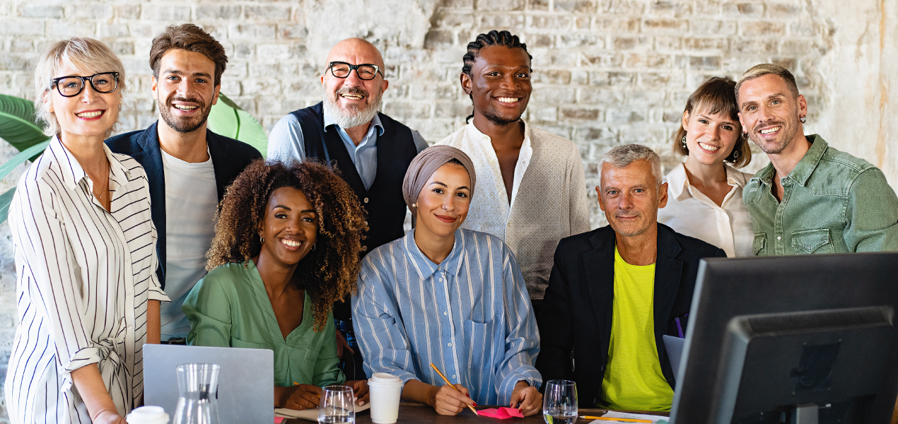 A diverse group of smiling professionals gathered in a modern office, collaborating around a computer.