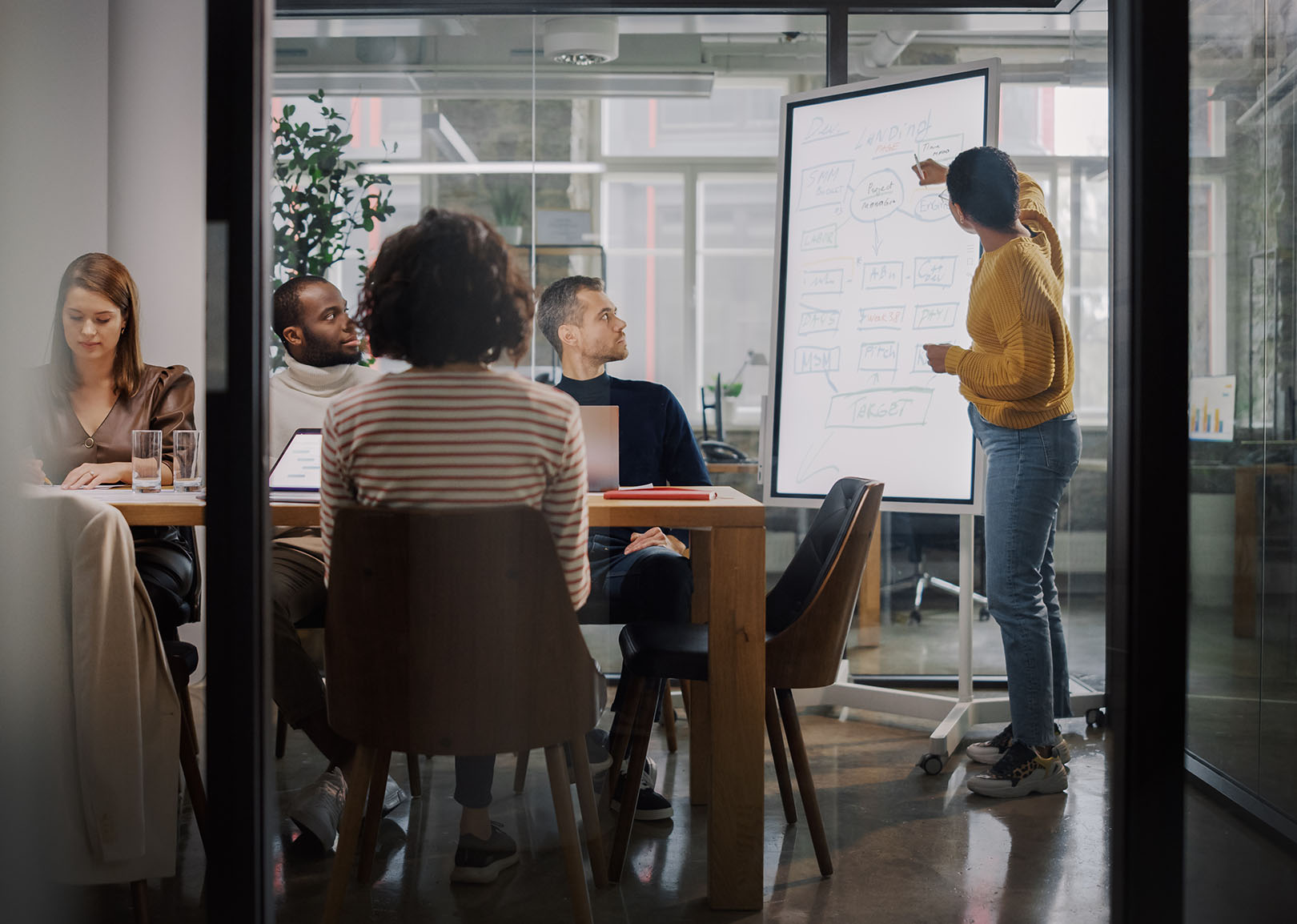 Team collaborating in a modern office, with a person presenting on a whiteboard and others seated around a table with laptops and notes.