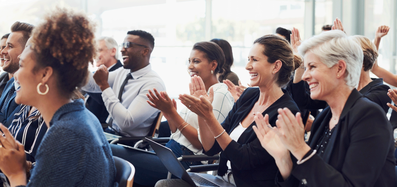 Large audience attending a professional conference, with people seated at tables and engaging in discussion in a brightly lit event space.