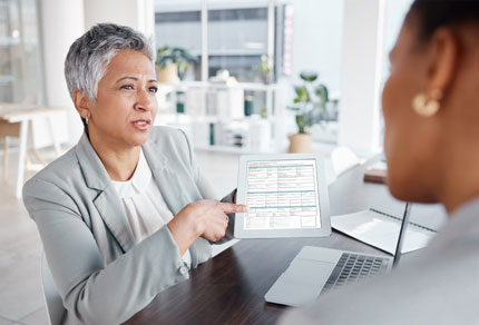 Businesswoman in a suit holding a document while offering advice, sitting at a desk with a blurred background.