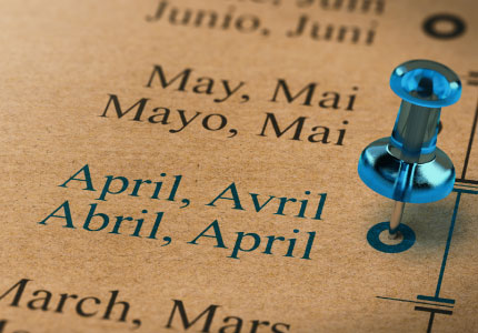 A close-up of a calendar or timeline showing the months "March" and "April" in multiple languages, with a blue pushpin marking a specific point on the timeline