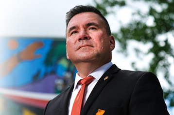 Bradley Regehr CBAPresident in 2020-21 wearing a suit and orange tie standing outdoors, with a mural and trees in the background.