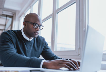 Businessman in a suit typing on a laptop at a desk with a blurred office background.