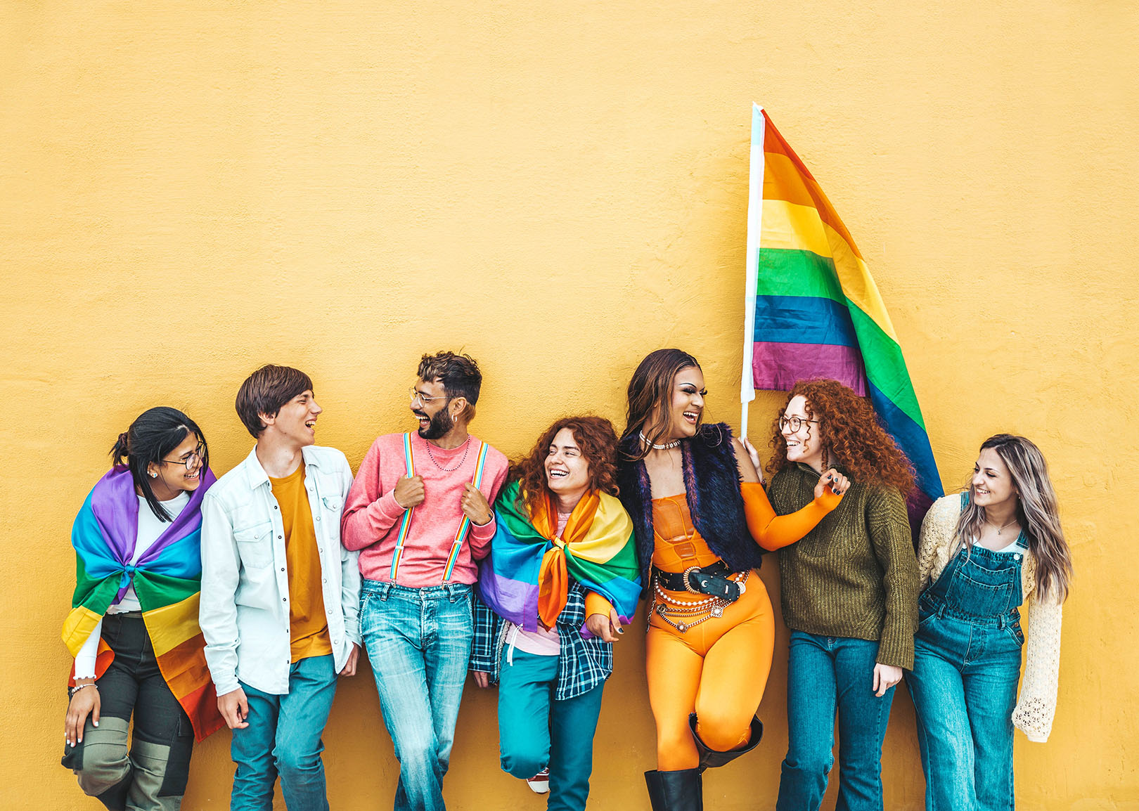 Group of diverse people smiling and standing against a yellow wall, with one person holding a rainbow Pride flag.