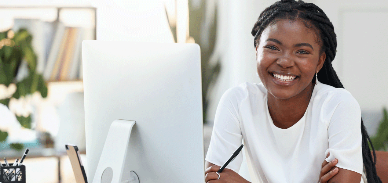 Smiling woman sitting at a desk in a bright, modern office, looking at a computer screen with a welcoming expression.