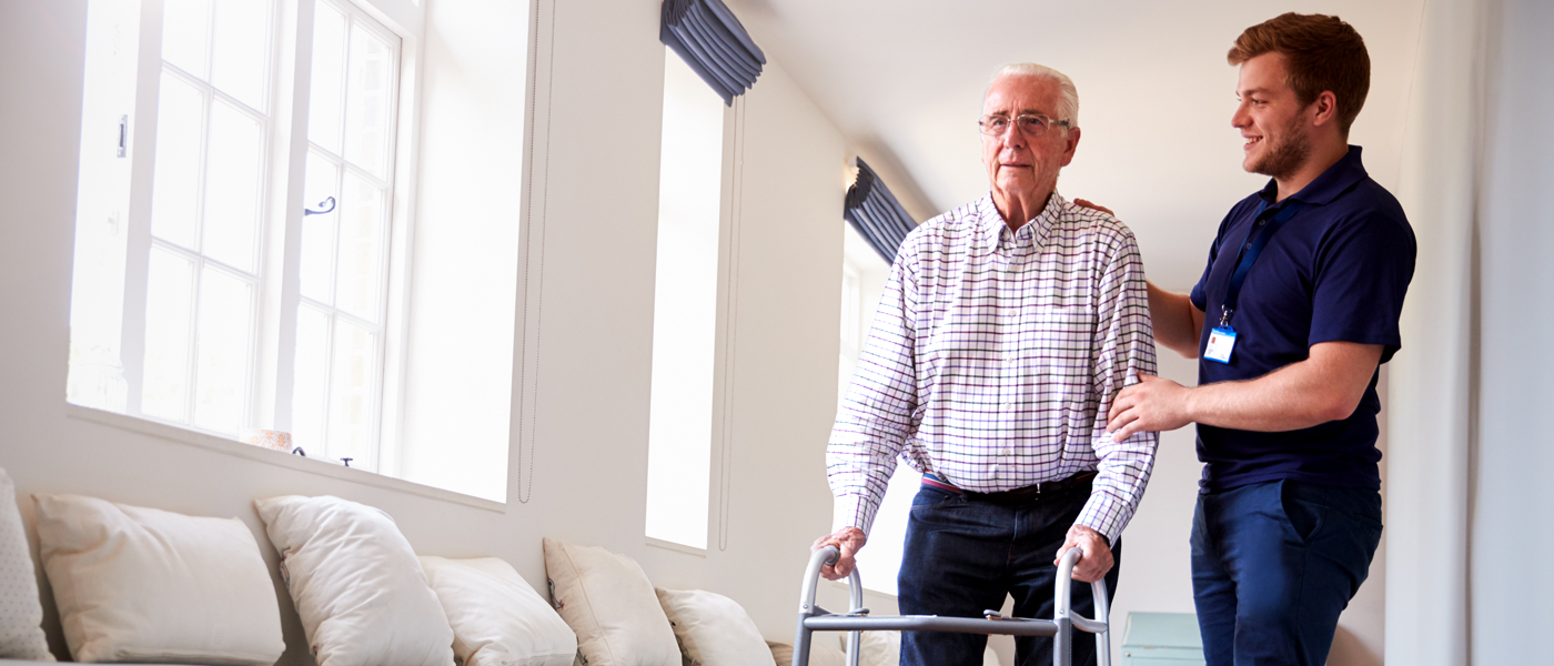 A healthcare worker assisting an elderly man using a walker in a bright medical or rehabilitation facility.