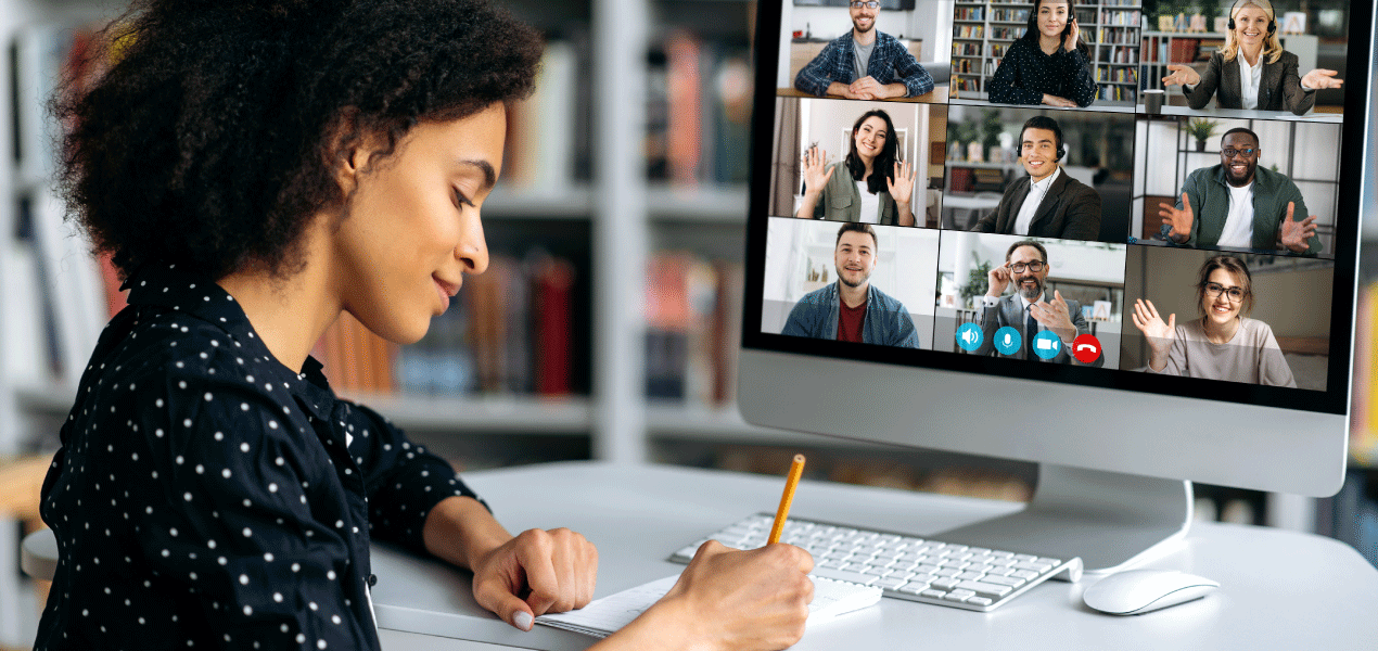 Person taking notes while participating in a virtual video conference on a desktop computer with multiple participants on the screen.