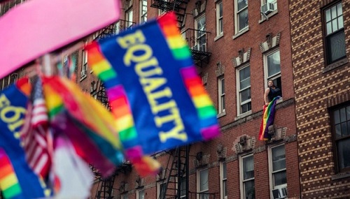 Equality rainbow flag in foreground with apartment building behind and person hanging out window waving their own flag.