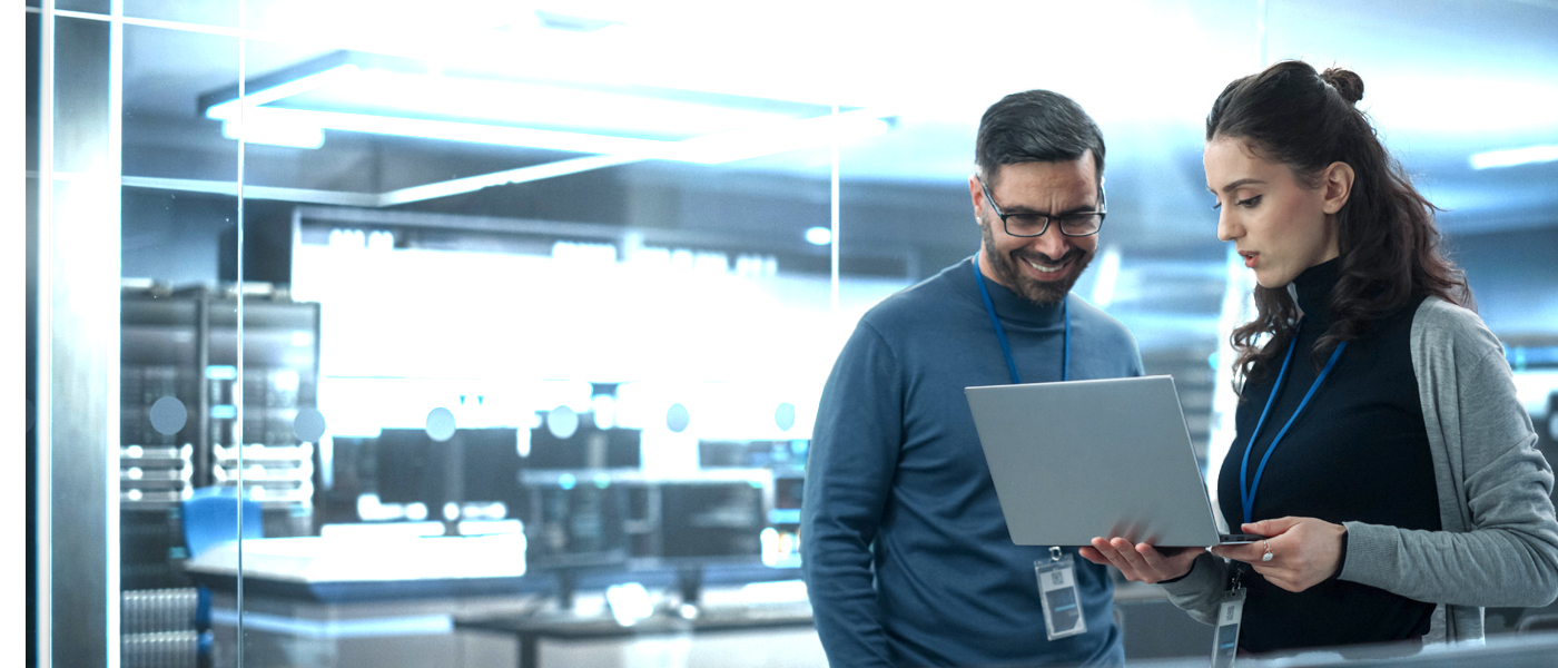 Two professionals collaborating in a high-tech laboratory or industrial setting, reviewing information on a laptop.