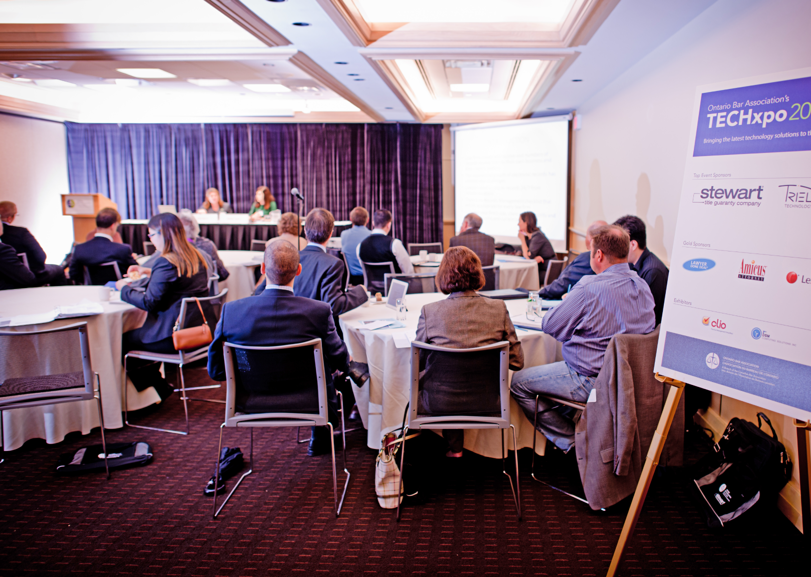  A professional conference setting featuring attendees seated at round tables, a panel of speakers at the front, and a branded event sign highlighting sponsors.