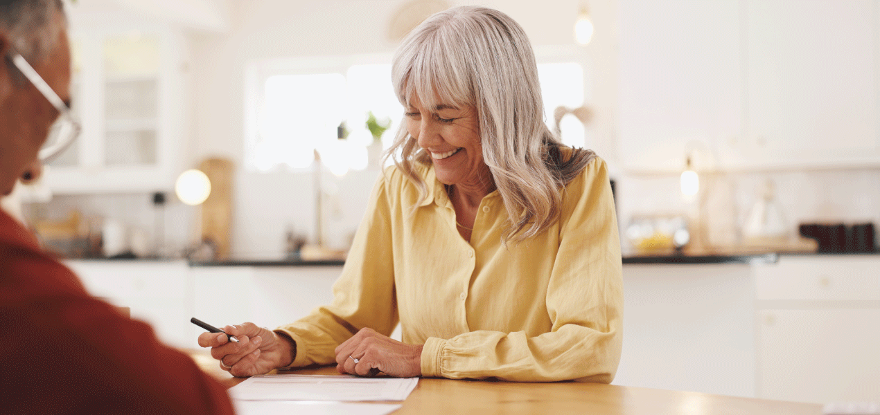 Smiling woman in a yellow shirt signing paperwork at a kitchen table in a bright home, suggesting will or estate planning.