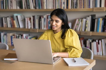 Woman in a yellow top working on a laptop at a library table, with shelves of books in the background.