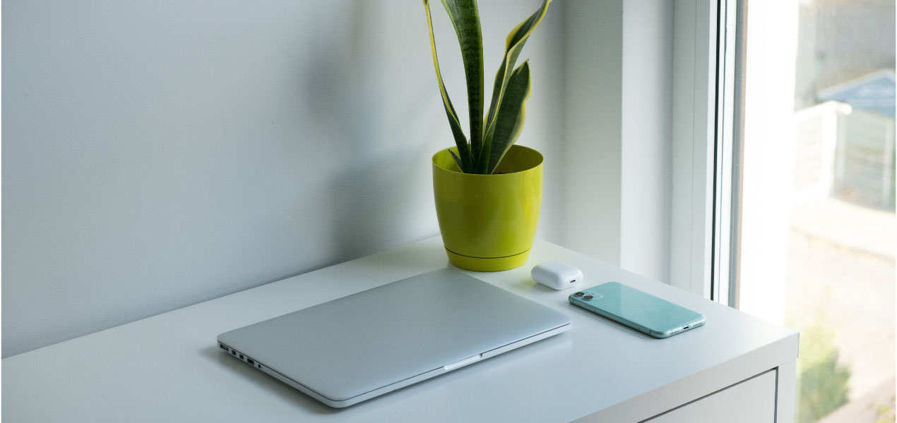 A closed laptop and a smartphone resting on a white desk near a window, with a green potted plant beside them, creating a clean and minimal workspace.