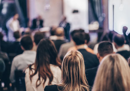 Audience attending a professional conference or seminar, focusing on speakers at the front of the room.