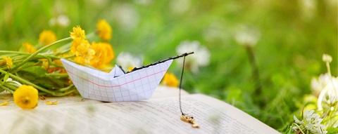 A paper boat made from graph paper placed on an open book, surrounded by yellow flowers and green grass in a serene outdoor setting.