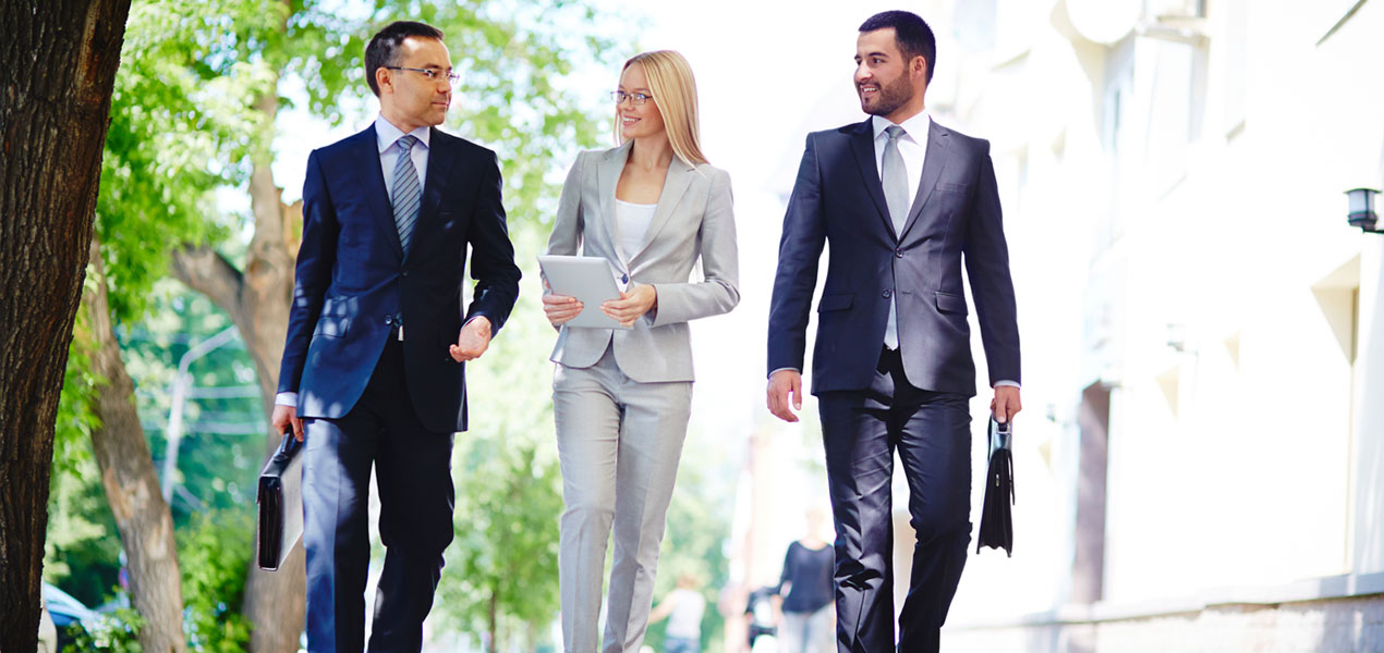 Three colleagues in business attire carrying briefcases are walking and talking outdoors.