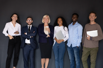 Group of six diverse professionals smiling and standing against a dark wall, holding devices and documents.