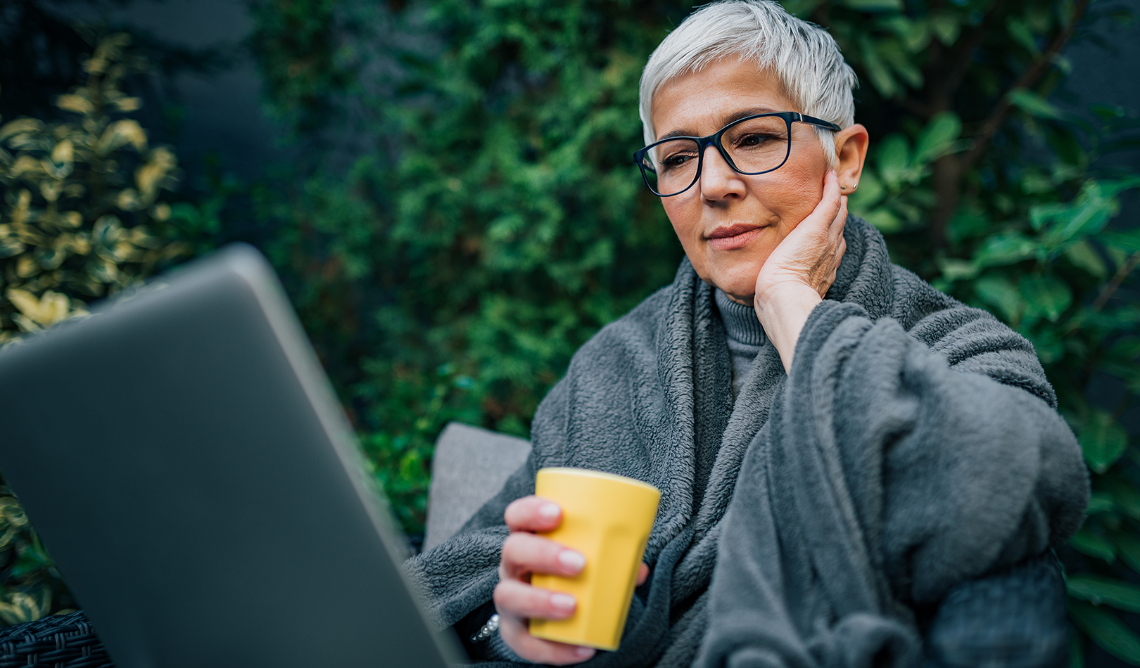 An older woman with short gray hair and glasses, wrapped in a gray blanket, sitting outdoors with a laptop and holding a yellow mug, looking focused.