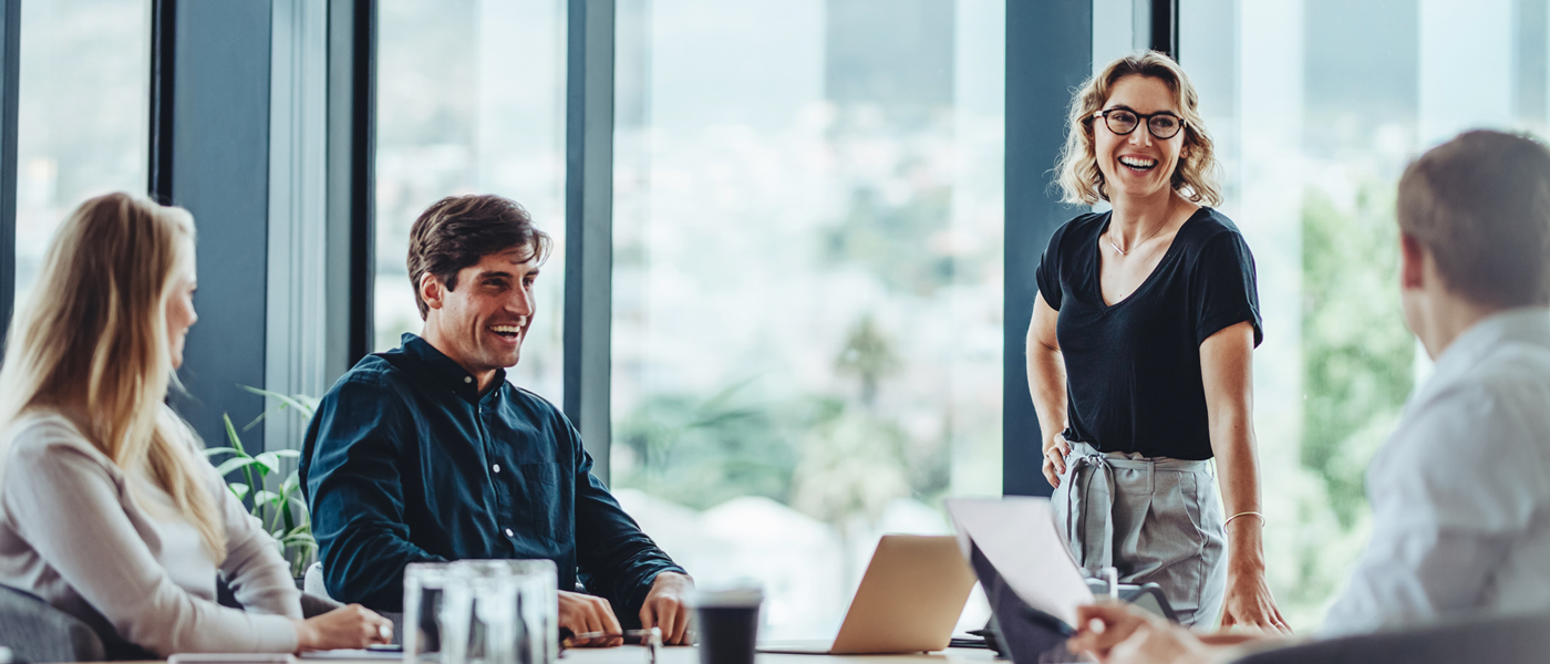 A group of professionals in a modern office engaged in a discussion, with a woman standing and presenting.