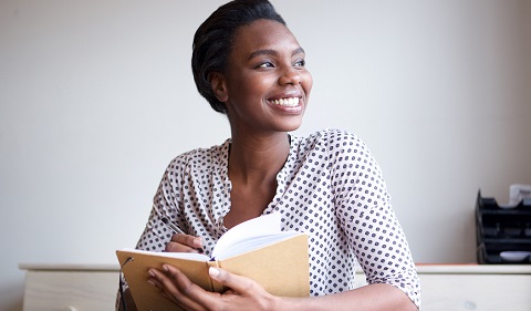 A smiling woman holding an open notebook, looking away thoughtfully, seated in a bright room with a neutral background.