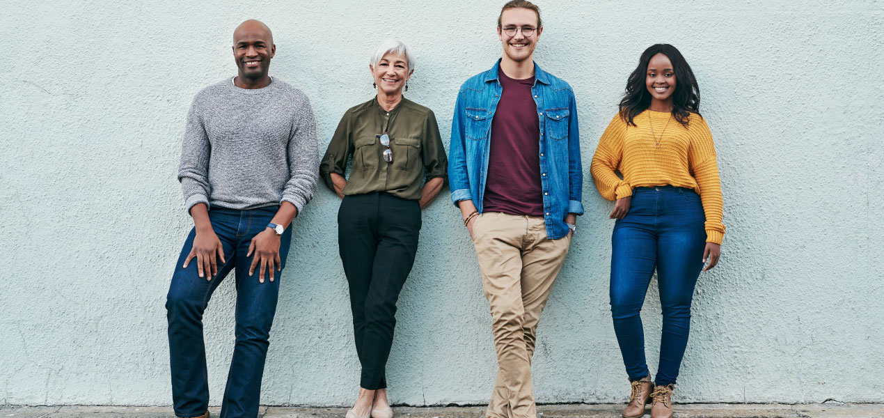 A diverse group of four casually dressed individuals standing against a light-colored wall, smiling confidently.