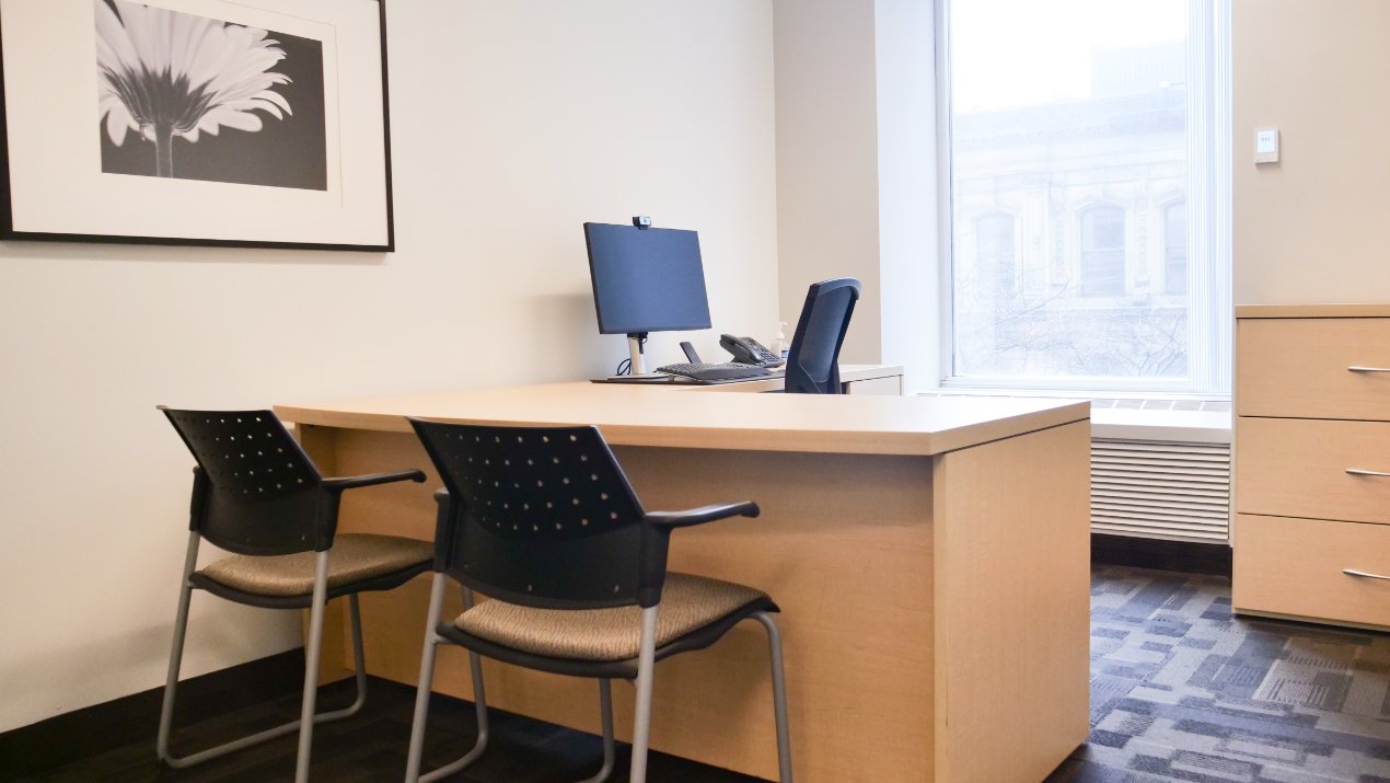 A modern and minimalistic office space featuring a wooden desk with two chairs, a computer, and a telephone, set against a bright window and neutral decor.