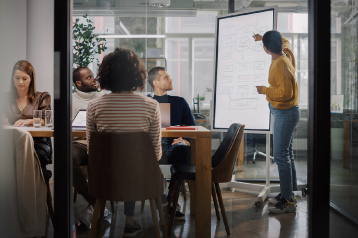 Team of people in a modern office meeting, with one person presenting ideas on a whiteboard while others sit at a table with laptops and notes.