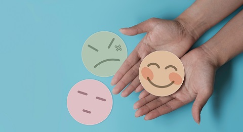 Hands holding a smiling face emoji cutout, with two other emoji cutouts (neutral and angry expressions) placed on a blue background.