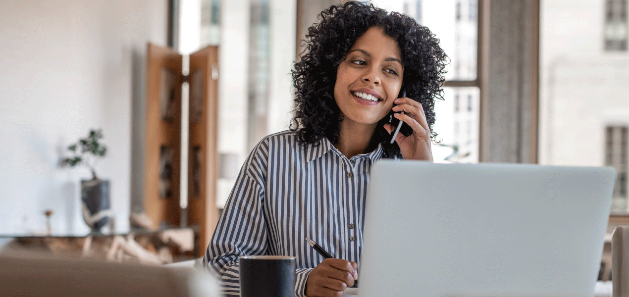 A smiling woman with curly hair working from home, talking on the phone while using a laptop. She is sitting at a desk with a coffee mug, in a bright and modern home office setting.