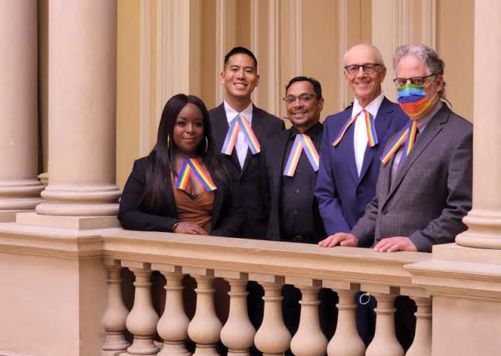 A group of five people wearing formal attire and rainbow tabs, standing in front of a balustrade with decorative columns in a bright indoor setting.