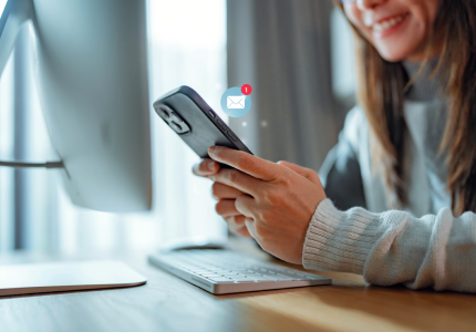 Woman smiling while checking her phone at a desk with a computer.