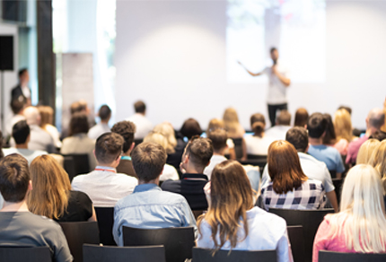 Audience seated at a professional conference listening to a speaker giving a presentation at the front of a modern meeting room.