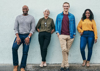 A diverse group of four casually dressed individuals standing against a light-colored wall, smiling confidently.