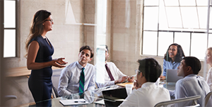 A professional woman standing and presenting to a group of business colleagues in a modern office meeting room.