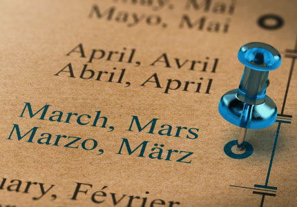 A close-up of a calendar or timeline showing the months "March" and "April" in multiple languages, with a blue pushpin marking a specific point on the timeline