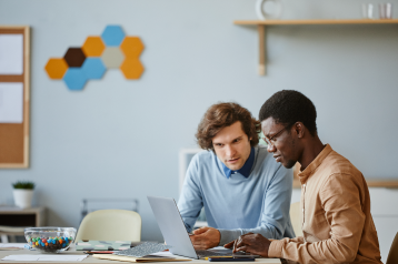 Two men collaborating at a desk, looking at a laptop, in a modern office setting with a corkboard and hexagonal wall art in the background.