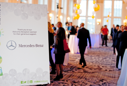 Event attendees networking in a formal venue with a sign thanking Mercedes-Benz as a sponsor in the foreground.