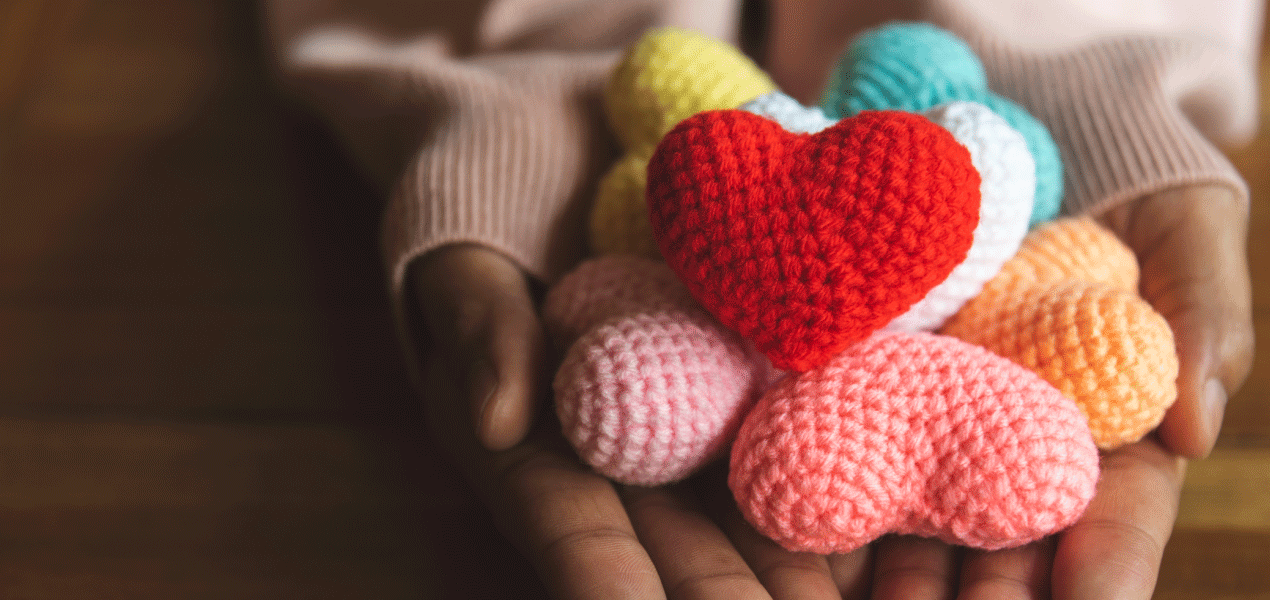 Hands gently holding a collection of colorful, knitted heart shapes, symbolizing warmth, kindness, and compassion.