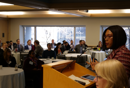 Woman speaking at a podium during an OBA Council meeting with an audience seated at round tables.