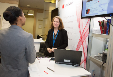 Two people conversing at a LexisNexis exhibit booth, featuring a laptop, promotional materials, and a display screen.