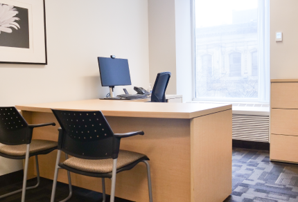 Minimalist office space with a wooden desk, two guest chairs, a computer monitor, and a large window providing natural light.