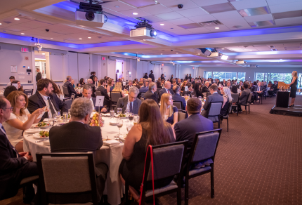 Banquet hall filled with formally dressed guests seated at round tables, enjoying a meal while listening to a speaker on stage.