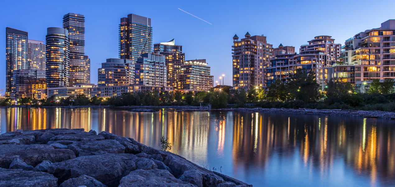 Scenic view of a modern city skyline at dusk, with illuminated buildings reflecting on a calm river and a rocky shoreline in the foreground.