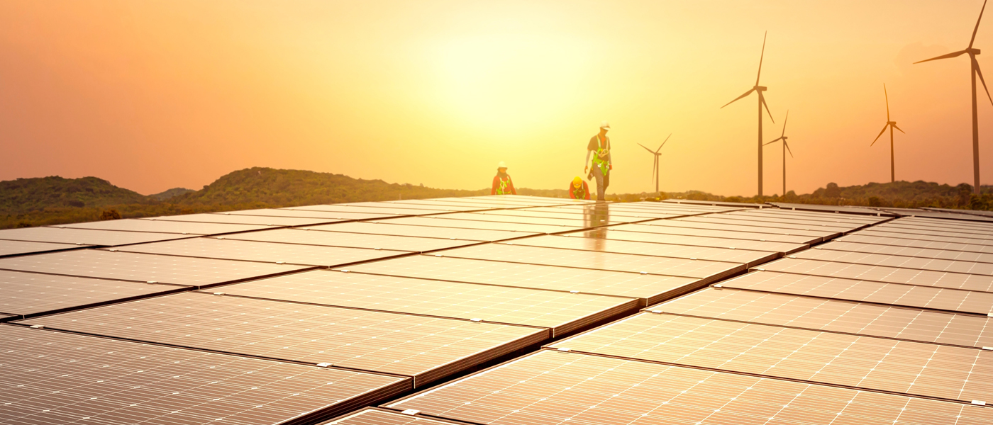 Solar panels installed in a field at sunset, with wind turbines in the background, representing renewable energy.