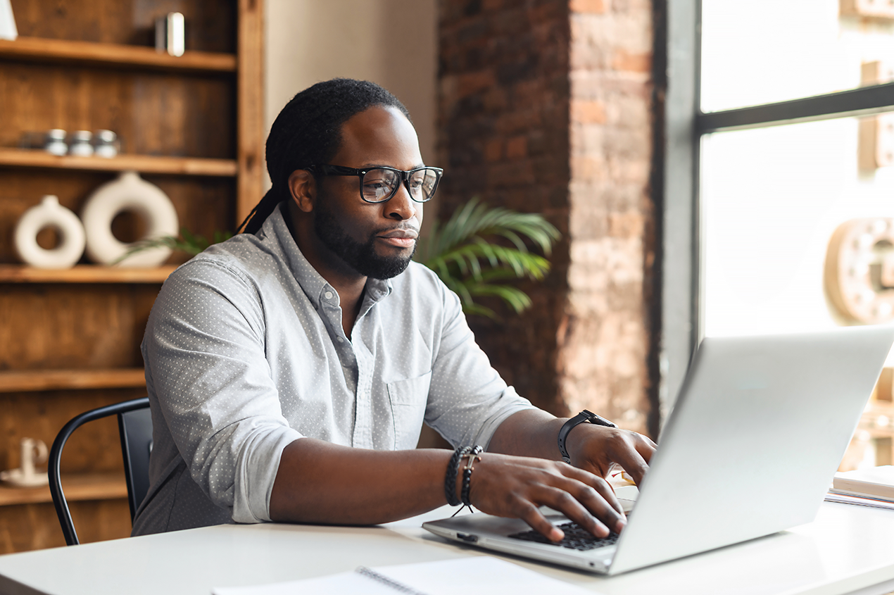 Man working on a laptop in a modern office setting with shelves and decorative items in the background.