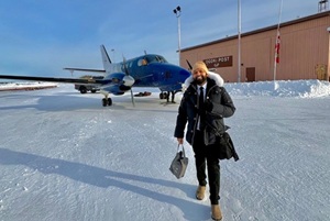 The author, in business suit, toque and parka, standing in front of small plane at airport, smiling, under blue skies